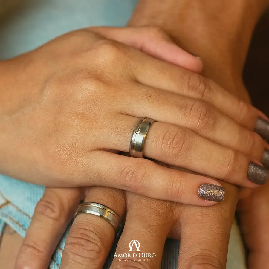 Par de anéis de compromisso com friso dourado e ponto de luz em mãos de casal