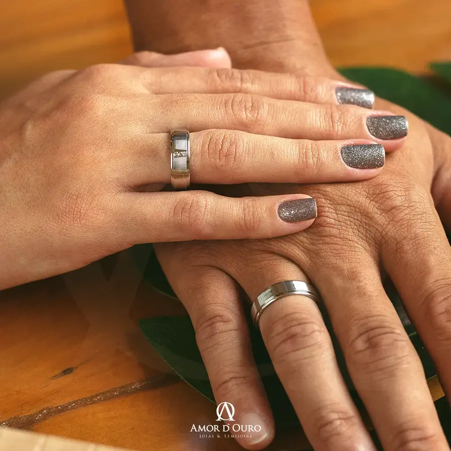 Par de anéis de compromisso prateados em mãos de casal sobre mesa de madeira